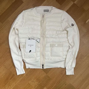 Vit Moncler dam-cardigan   - Snygg vit dam-cardigan jacka från Moncler med quiltad front och två fickor. Jackan har ribbade muddar, dragkedja framtill och Moncler-logga på ärmen. Ärmarna är stickade och kroppen är i ett glansigt, lätt vadderat material. Perfekt för lager-på-lager-stil. Helt nytt skick
