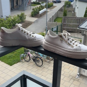 Vita sneakers från Rieker med dragkedja - Snygga vita sneakers från Rieker med chunky sula, guldfärgade detaljer och dragkedja på sidan. Skorna har snörning och är gjorda i skinn med robust platt sula. Perfekta för dig som gillar stilrena och trendiga skor med en twist.