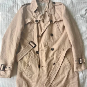 Beige trenchcoat från Kiomi - Stilren beige trenchcoat från Kiomi med klassisk dubbelknäppning, axelklaffar och bälte i midjan. Jackan har långa ärmar, stora fickor och snygga spännen vid ärmslut och midja. Perfekt för dig som vill ha en tidlös look.