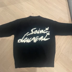 Svart stickad Saint Laurent tröja - Svart stickad tröja från Saint Laurent med vit, stor logga i graffiti-stil över bröstet. Tröjan har rund halsringning, långa ärmar och ribbade muddar. Perfekt för dig som vill ha en statement piece med lyxig känsla och street vibe. Storlek S och väldigt fint skick. Priset inte hugget i sten!