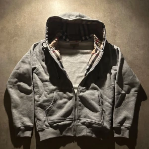 Grå kofta från Burberry - Grå kofta från Burberry med dragkedja och klassiskt rutigt foder i huvan. Koftan har två stora fickor framtill, ribbade muddar och är tillverkad i mjuk bomull. Perfekt för dig som vill ha en stilren och bekväm look med en touch av lyx. Hör av vid minsta fundering 💫