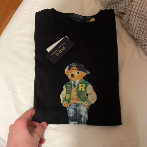 Svart Polo Bear t-shirt från Ralph Lauren - Svart t-shirt från Polo Ralph Lauren med klassisk passform. Framsidan har en stor Polo Bear-tryck där björnen bär collegejacka, jeans och sneakers. Rund halsringning och korta ärmar. Tillverkad i mjuk bomull för skön känsla.