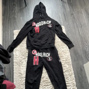 Svart HOODRICH set  - Svart set från HOODRICH med stor vit och röd logga och patchar på bröstet. Tröjan har huva med snörning och långa ärmar. Materialet är mjukt och fleeceliknande på insidan, perfekt för chill dagar. Strl S 