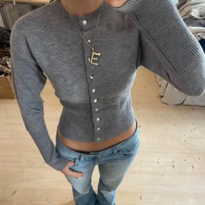 Grå croppad kofta med knappar - Supersnygg grå croppad kofta med långa ärmar och knappar framtill. Tröjan har ribbade muddar och midja som ger en tight och figurnära passform. Perfekt att styla med jeans för en chill look.