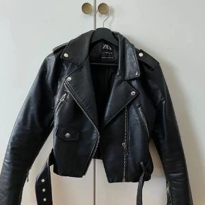 Svart skinnjacka från Zara med bälte - Snygg svart skinnjacka från Zara med coola detaljer som dragkedjor, axelklaffar och bälte i midjan. Jackan är croppad och har klassisk bikerstil med silvriga knappar och dragkedjor. Perfekt för dig som vill ha en edgy look. Endast använd 2-3 ggr och är nu inte min stil💐