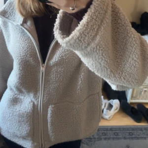 Beige teddyjacka med dragkedja - Supermjuk och fluffig beige teddyjacka med hel dragkedja framtill och stora fickor. Jackan har en avslappnad passform och är perfekt för kyliga dagar. Den är långärmad och har en mysig känsla som passar till alla outfits.