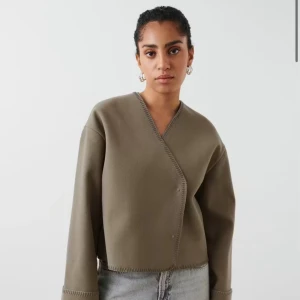 Beige omlott overshirt med knappar - Säljer en beige omlottjacka i overshirt-modell med v-ringning och dekorativa knappar framtill. Jackan har lång ärm, croppad passform och synliga sömmar längs kanterna för en clean look. Perfekt att slänga över en t-shirt eller linne. Använd någon gång och är super skön nu i höst😍Skriv om egna bilder 