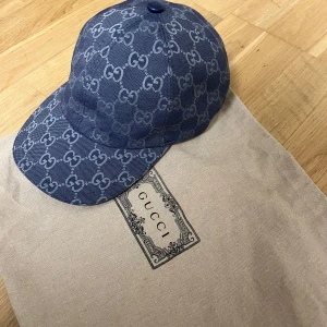 Blå keps från Gucci med GG-mönster - Snygg blå keps från Gucci med ikoniskt GG-monogram över hela kepsen. Tillverkad i bomullstyg med klassisk böjd skärm och justerbar baksida. Perfekt accessoar för att lyfta din outfit med lyxig streetstyle-vibe.