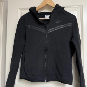 Svart Nike Tech Hoodie - Svart Nike Tech hoodiejacka med hel dragkedja och luva. Snygga svarta detaljer över bröstet och diskret Nike-logga. Jackan har långa ärmar och två fickor framtill. Perfekt för chill dagar eller när du vill ha en sportig look.