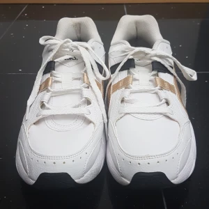 Vita Asics sneakers med gulddetaljer - Snygga chunky Asics sneakers i vitt med guld- och svarta detaljer. Skorna har klassisk snörning, rund tå och platt sula. Ovandelen är i syntetmaterial och de har en sportig vibe med GEL-dämpning i sulan. Perfekta för dig som gillar streetstyle och bekväma skor. PRIS KAN DISKUTERAS!