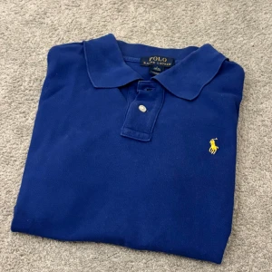 Blå pikétröja från Ralph Lauren - Klassisk blå pikétröja från Ralph Lauren med gul broderad logga på bröstet. Tröjan har krage, två knappar och korta ärmar. Tillverkad i bomull och har en snygg, tidlös look.