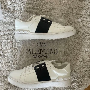 Valentino Garavani sneakers med svart detalj - Snygga vita sneakers från Valentino Garavani med en bred svart rem över sidan och ikoniska nitar på sulan. Skorna har snörning och en klassisk rund tå. Perfekta för dig som vill ha en stilren och exklusiv look.