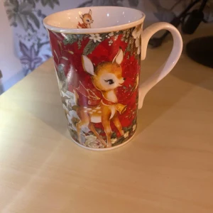 Julkopp med rådjur och blommor - Säljer en söt porslinskopp med julmotiv. Koppen har ett rött mönster med ett tecknat rådjur, vita blommor och gröna blad. Insidan har också en liten bild på rådjuret. Perfekt för varma drycker under vintern.