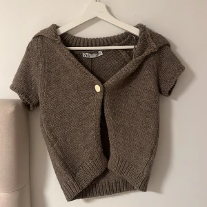 Brun stickad kofta med kort ärm från Zara - Säljer en brun stickad kofta från Zara med korta ärmar. Koftan har en knapp framtill och ribbade kanter. Perfekt för lager-på-lager och enkel att styla till olika outfits. Den är oanvänd och har även lappen kvar på sig. 