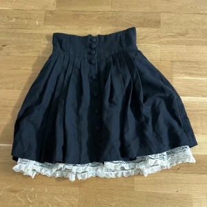 Svart kjol med knappar och spetskant - Säljer en svart kjol med hög midja och knappar framtill. Från H&M och går till knäna. Kjolen har veckad design och en vit spetskant som sticker ut nedtill för en extra detalj. Storlek 34!