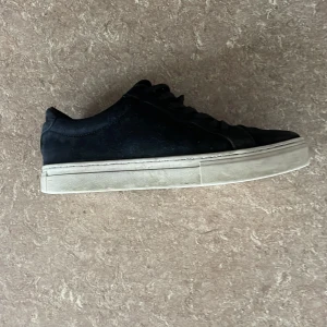 Svarta låga sneakers - Säljer ett par svarta sneakers med platt vit sula och rund tå. Skorna har snörning och är tillverkade i mocka. Perfekta för en stilren och enkel look.