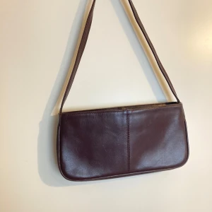 Brun axelväska i skinn - Säljer en stilren brun axelväska i skinn med enkel design och raka linjer. Väskan har ett  axelband och stängs med knapp, samt ett litet fack med dragkedja. 