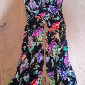 Blommiga byxor från H&M storlek 34 - Säljer färgglada byxor från H&M med svart botten och stora blommor i lila, rött, grönt och blått. De har en flowy passform och är perfekt för sommarens tillfällen. De har en lite satin look. Använda enstaka gång på grund av för små. Inga defekter 