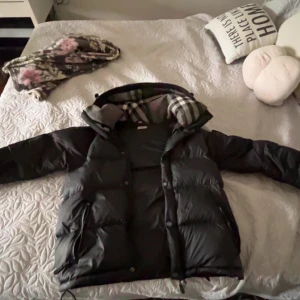 Burberry jacka  - Säljer en svart dunjacka från Burberry med klassiskt rutigt foder i huvan. Jackan har lång ärm, dragkedja och knappar framtill. Perfekt för kyliga dagar och har en stilren design med hög krage. Obs dessvärre så är vänster dragkedja trasig men är möjligt att laga för någon hundra lapp