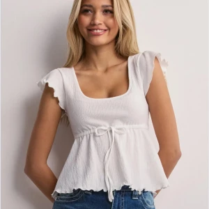 Vit topp Nelly - Topp från Nelly ”Cute frill tie top”, köpt förra sommaren och använd endast några gånger. Storlek XS men passar även mig som har S. Mycket bra skick, inget att anmärka på.