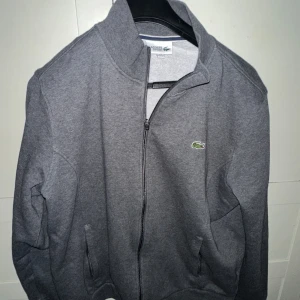 Grå zip-jacka från Lacoste - Säljer en stilren grå jacka från Lacoste med dragkedja framtill och klassisk krokodil-logga på bröstet. Jackan har hög krage och två fickor med dragkedja. Perfekt för dig som gillar enkel och sportig stil.