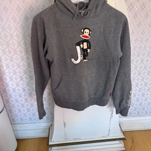Grå hoodie från Paul Frank med apa-tryck - Säljer en grå hoodie från Paul Frank med det klassiska apa-trycket på bröstet och texten 'julius' på ärmen. Tröjan har huva med dragsko och en stor magficka. Perfekt för dig som gillar streetwear och vill ha något bekvämt och snyggt.