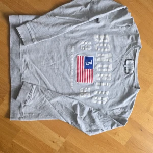 Ljusgrå långärmad tröja  - Ljusgrå långärmad tröja  med tryckt text och amerikansk flagga på bröstet. Tröjan har rund halsringning och ribbade muddar vid ärmslut och nederkant. Perfekt för en avslappnad stil.