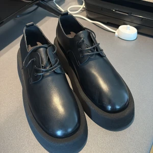 Svarta Marsèll platåskor med snörning från H&M - Säljer ett par svarta skorfrån Marsèll med snörning. Skorna har en rund tå och en kraftig platt sula som ger extra höjd. Tillverkade av riktig djur skinn perfekta för dig som vill sticka ut lite extra. Helt nytt skick. 