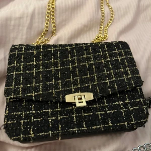 Svart och guld axelväska med kedja - Säljer en snygg svart axelväska med guldiga detaljer och kedjerem. Väskan har ett rutigt mönster i svart och guld samt ett elegant spänne framtill. Perfekt för att lyfta din outfit med lite extra stil. Passar perfekt att ha till festliga tillfällen men även när man vill pimpa till vardagen!! Väskan är i bra skick trots att den använts en del.