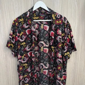 Svart blommig skjorta från Brave Soul - Säljer en svart skjorta med färgglatt blommigt mönster i rött, gult och vitt från Brave Soul. Skjortan har korta ärmar och en avslappnad passform, perfekt för sommarens alla tillfällen.