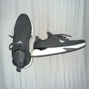 Svarta sneakers från Emporio Armani - Säljer ett par svarta sneakers från Emporio Armani med vit sula och stickad ovandel. Skorna har snörning och logga på sidan samt platt sula för en clean look.