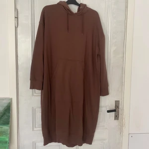 Brun hoodieklänning från 157 - Mysig brun hoodieklänning från 157 med huva och dragsko. Klänningen har lång ärm, stor känguruficka framtill och avslappnad passform. Perfekt för en chill och bekväm stil.