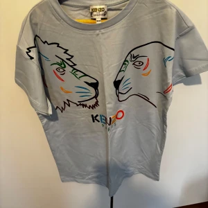 Ljusblå t-shirt med tryck från Kenzo Kids - Säljer en ljusblå t-shirt från Kenzo Kids med färgglatt tryck av två djurhuvuden på framsidan och Kenzo Paris-logga. T-shirten är i mjuk bomull och har en klassisk passform med korta ärmar. Perfekt för dig som gillar unika prints! Aldrig använd, prislappen borttagen dock!
