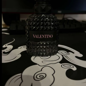 Svart parfymflaska från Valentino - Säljer en lyxig parfym från Valentino i en svart, mönstrad flaska med nitar och elegant etikett. Flaskan har en rundad form och ger ett exklusivt intryck, perfekt för dig som gillar stilrena detaljer.