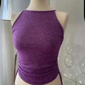 Lila glittrig croppad linnetopp S/M - Säljer en lila glittrig croppad linnetopp med smala axelband och åtsittande passform. Toppen har en snygg glansig yta och är perfekt för dig som vill sticka ut lite extra.