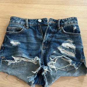 Blå slitna jeansshorts med fransar - Säljer ett par blå jeansshorts med slitna detaljer och fransar nertill. Shortsen har klassisk femficksdesign, knapp och dragkedja framtill samt råa kanter. Perfekta för en avslappnad stil.