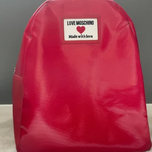 Ryggsäck Moschino  - En superfin och sjävklart äkta ryggsäck i konstläder från det italienska lyxmärket Moschino. Storlek 26x31x14cm. Väskan är helt ny alltså den har aldrig använts. Nypris ca 1850 sek. 