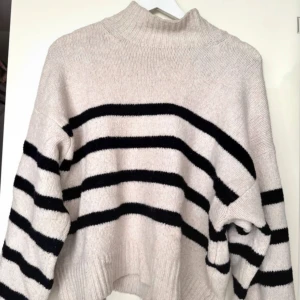 Beige och svart randig stickad tröja - Säljer en mysig stickad tröja i beige med svarta ränder. Tröjan har hög krage, långa ärmar och en något croppad passform. Perfekt för kyliga dagar och enkel att styla till jeans eller kjol.