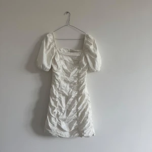 Vit klänning från NA-KD - Säljer en vit kortklänning från Chloé B x NA-KD i storlek 34. Klänningen har puffärmar, fyrkantig ringning och rynkade detaljer längs sidorna. Perfekt för sommarens tillfällen, studenten eller andra roliga händelser! 