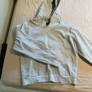 Ljusgrå hoodie från 157 - Säljer en enkel och stilren ljusgrå hoodie från 157. Tröjan har huva med dragsko och en stor magficka framtill. Den är klippt vid halsen eftersom den satt för tight