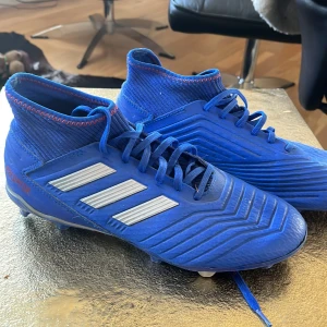 Blå Adidas Predator fotbollsskor - Säljer ett par blå Adidas Predator fotbollsskor med vita ränder och röda detaljer. Skorna har snörning, hög krage i stickad syntet och mönstrad ovandel för extra grepp. Perfekta för spel på gräsplaner.