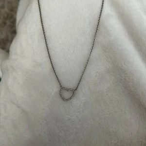 Halsband med hjärthänge i silverfärg - Säljer ett fint halsband i silverfärg med ett hjärthänge dekorerat med små glittrande stenar. Perfekt accessoar för att lyfta din outfit med en söt detalj. Kedjan är tunn och smidig.