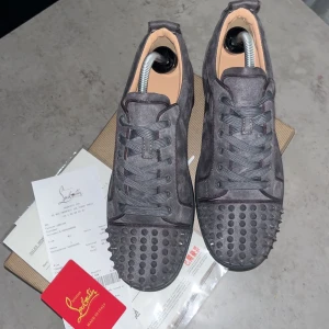 Grå sneakers med nitar från Christian Louboutin - Säljer ett par grå sneakers från Christian Louboutin med ikonisk röd sula och nitar framtill. Skorna har snörning och är tillverkade i ett lyxigt material med en unik design som sticker ut.