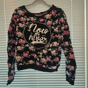Svart blommig sweatshirt från Chaserdivas - Svart sweatshirt med färgglatt blommönster och vit text 'Now or never' på bröstet. Tröjan har rund halsringning, långa ärmar och ribbade muddar. Perfekt för dig som gillar en avslappnad men ändå snygg stil.