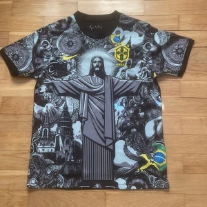 Brasilien fotbollströja med Kristusstatyn - Unik Brasilien fotbollströja från Nike med stort tryck av Kristusstatyn och brasilianska motiv. Tröjan är svart med detaljer i vitt, gult och blått samt har korta ärmar och rund hals. Perfekt för dig som vill sticka ut på läktaren eller i stan.
