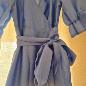 Blå omlottklänning med puffärm - Superfin ljusblå omlottklänning från Ginq Tricot med puffiga korta ärmar och bred knytskärp i midjan. Klänningen har v-ringning och faller i en elegant, vadlång siluett. Helt oanvänd säljer pga att den va för liten vid mina bröst och hann inte returnera. Superfin klänningen och underbar färg.