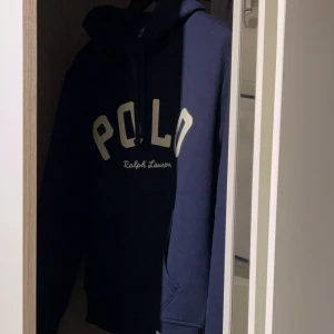 Mörkblå hoodie från Polo Ralph Lauren - Säljer en mörkblå hoodie från Polo Ralph Lauren med stor vit logga på bröstet och huva med dragsko. Klassisk och stilren modell som passar perfekt till vardags. Mjuk insida och bekväm passform. Storlek XS Passar runt 170, knappt använd. Nypris ligger på 2000:-