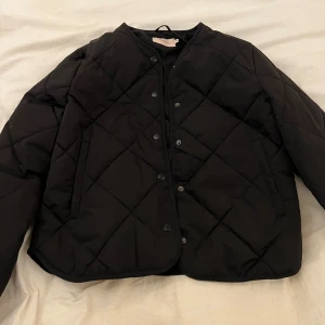 Svart quiltad jacka från Nelly - Säljer en svart quiltad jacka från Nelly i storlek M. Jackan har tryckknappar framtill, rund halsringning och två fickor. Perfekt för lager-på-lager och enkel att matcha till olika outfits.
