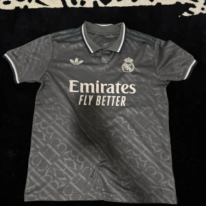 Real Madrid grå fotbollströja från Adidas - Säljer en grå Real Madrid fotbollströja från Adidas med klubbmärke och sponsortryck på bröstet. Tröjan har korta ärmar, krage med knapp och subtilt mönster i tyget. Perfekt för dig som vill visa ditt stöd för laget.