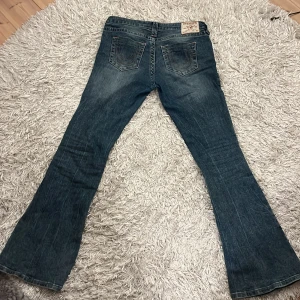 Blå bootcut jeans från True Religion - Säljer ett par klassiska blå bootcut jeans från True Religion med snygga kontrastsömmar och ikonisk bakficka. Jeansen har normal passform och är tillverkade i slitstarkt denim. Perfekta för dig som gillar en tidlös jeansstil.